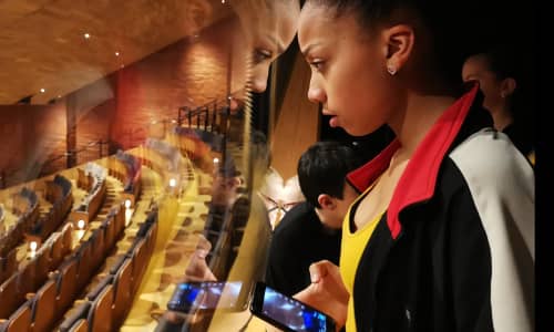 A young black girl holding a mobile phone looks through a window into an empty theatre