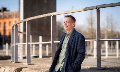 A man wearing a dark blue, long jacket, smiling and leaning against a wall