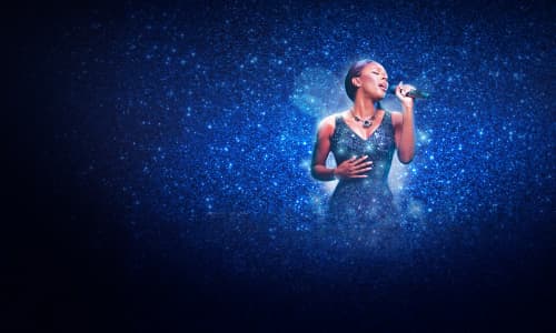 Alexandra Burke wearing a blue dress singing into a microphone on stage during The Bodyguard The Musical