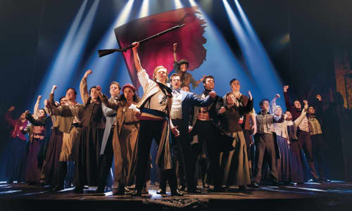 A scene from Les Mis featuring actors on stage holding banners and flags in a defiant manner.