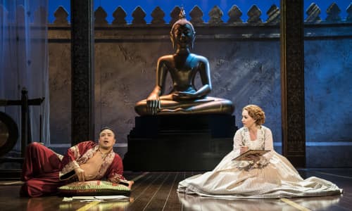 A scene from the King and I featuring a man dressed in oriental costume and a woman wearing a dress, sitting on the floor next to a statue