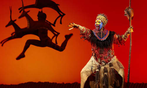 Scene from The Lion King featuring an orange background, gazelles leaping and an African chief with spear singing