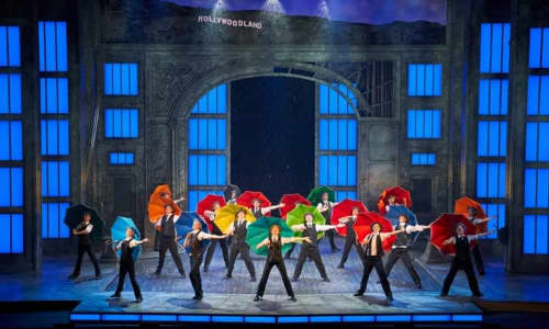 The cast of Singing In The Rain dancing with colourful umbrellas on a stage