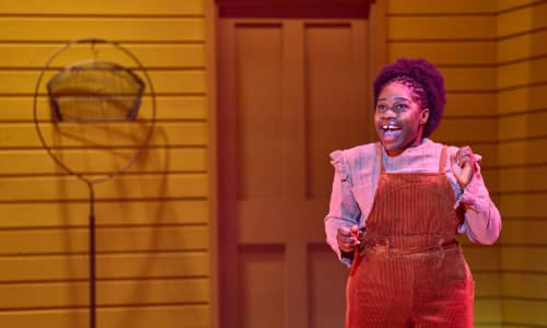 A Black woman singing on stage in brown corduroy dungarees