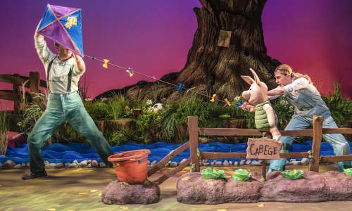 A puppet of Piglet flying a kite on stage, next to a cabbage patch, with puppeteers handling Piglet and the kite