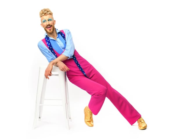 Andrew Pepper is dressed in a pink and blue outfit, leaning on a white stool.