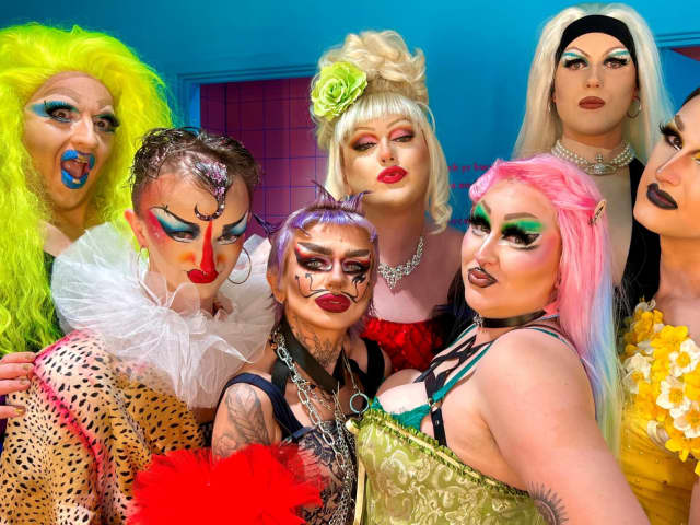 Seven drag performers in colourful clothes and makeup pout towards the camera.