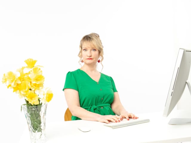 A woman sat at an office desk in a bright green dress with a vase of daffodils on the desk