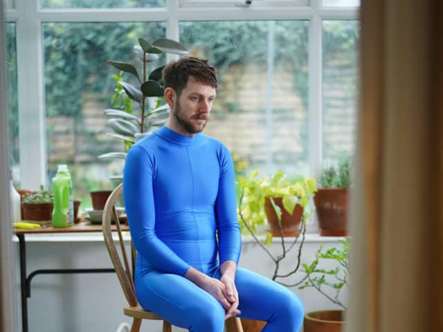 A person sat on a wooden chair in a conservatory dressed in a blue leotard