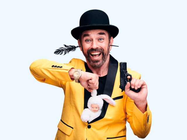 Kizza wears a bowler hat and bright yellow blazer holding a toy gun and toy baby in a rabbit suit.