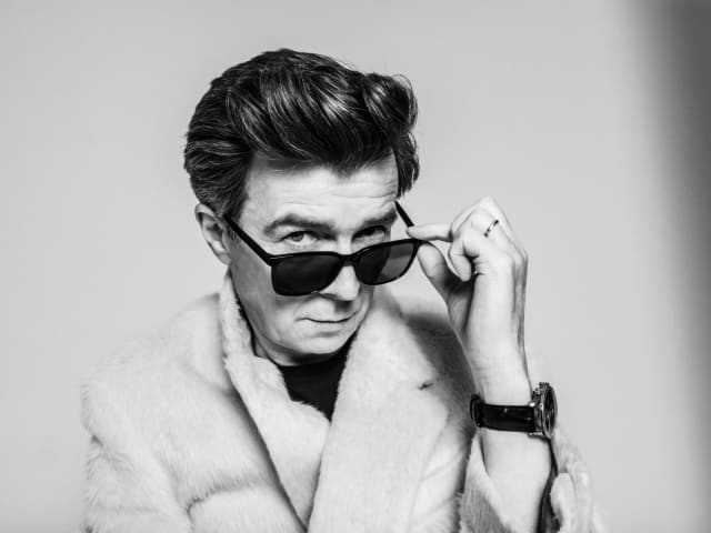 A black and white image of popstar Rick Astley peering over his sunglasses