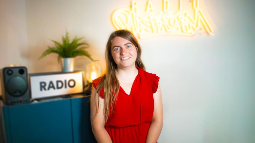 Agathe has long, light brown hair and is wearing a red dress. She is standing in the Radio Platfform studio.