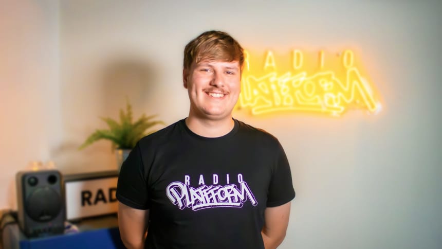 Dylan has short brown hair and a moustache. He is wearing a black Radio Platfform tshirt.