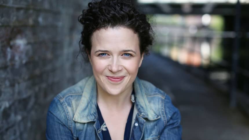 A white woman with dark curly hair wearing a denim jacket, smiling at the camera