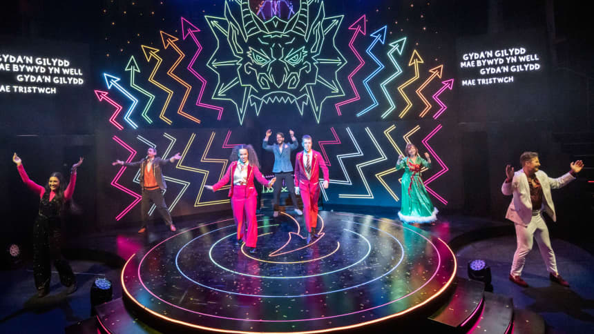 The TV talent show contestants on stage with a neon rainbow dragon in the background