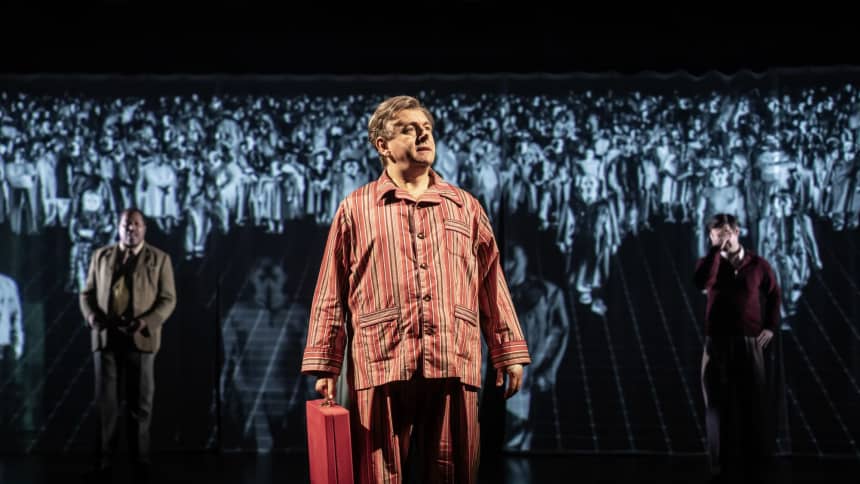 Michael Sheen as Nye Bevan, holding a ministerial red box with a crowd of people behind him projected onto a screen