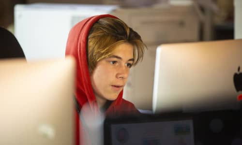 A teenage boy wearing a red hoodie sat at a computer screen