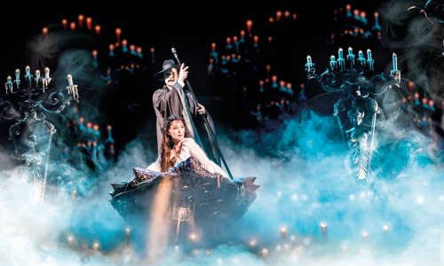 A man dressed as the Phantom steers a boat containing a woman through a misty scene with lights and candles either side of them.