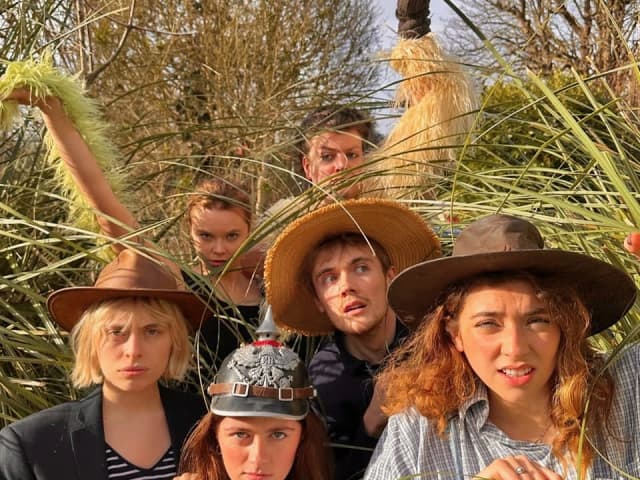 A group crouching in long grass wearing Australian-style sun hats, with two puppet emus behind them