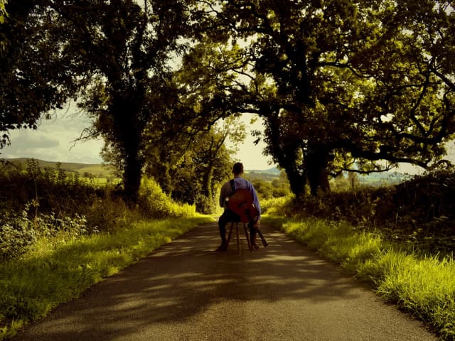 Vernon James sits on a stool in the middle a country road with his guitar and back towards us.