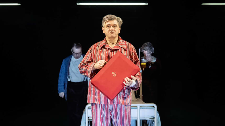 Michael Sheen dressed in striped pyjamas as Nye Bevan, holding a red ministerial box in front of a hospital bed