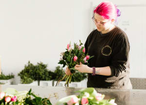 A person with pink hair and glasses stands arranging bouquets of fresh flowers