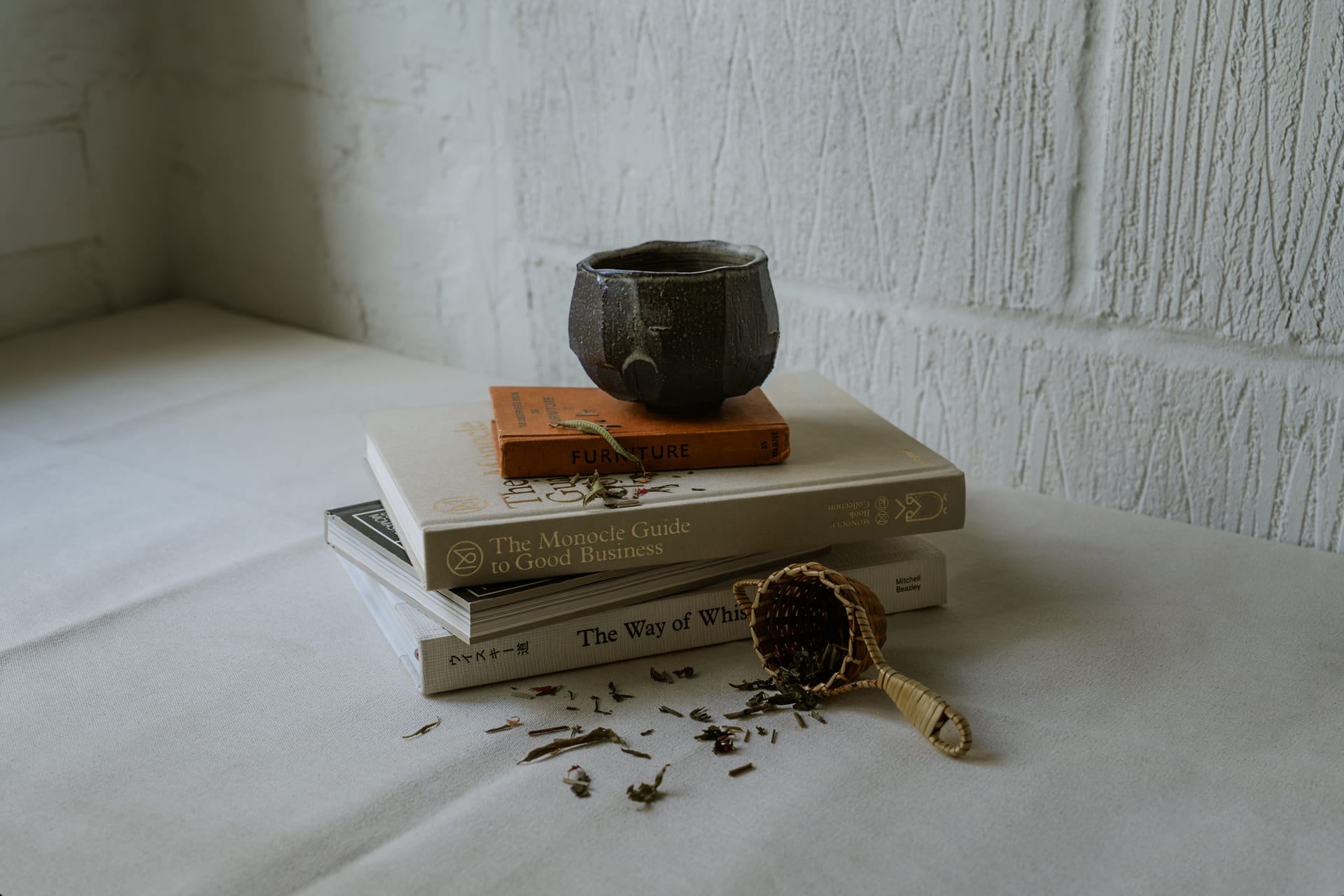 Japanese ceramic tea cup on top of books and magazines with tea leaves spread around and a white concrete wall in the background