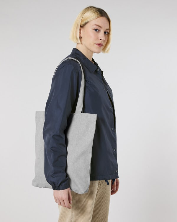 Person in navy blue jacket and beige trousers carrying light grey fabric tote bag, promoting eco-friendly accessories.