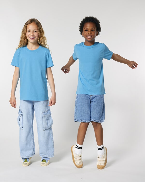 Two children wearing blue organic cotton t-shirts and denim bottoms, promoting eco-friendly kidswear on neutral backdrop