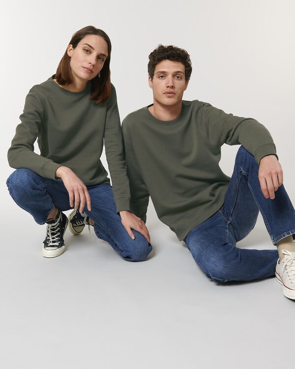 Two people in matching green organic sweatshirts and jeans, promoting sustainable fashion on a neutral background.