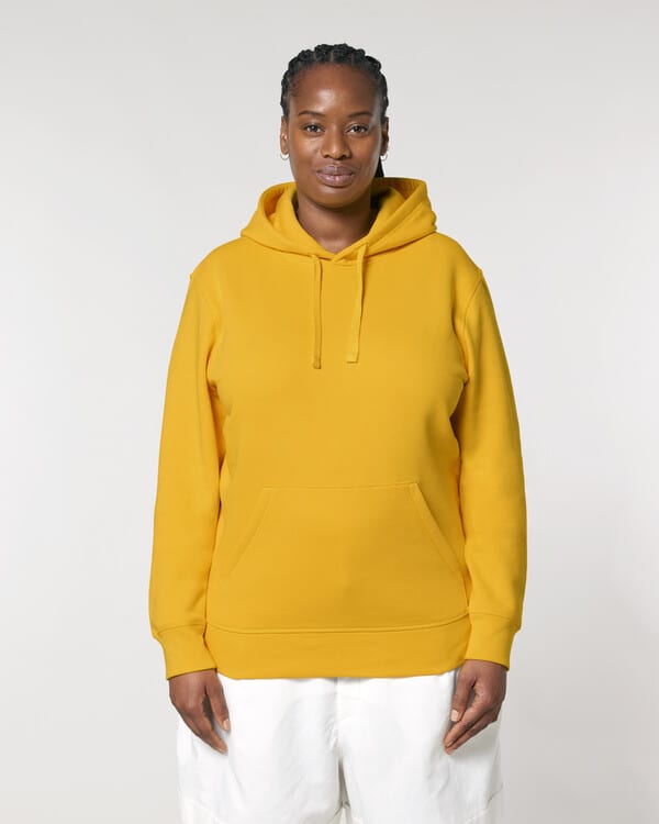 Woman in bright yellow organic hoodie and white trousers, showcasing ethical streetwear on a plain neutral background.