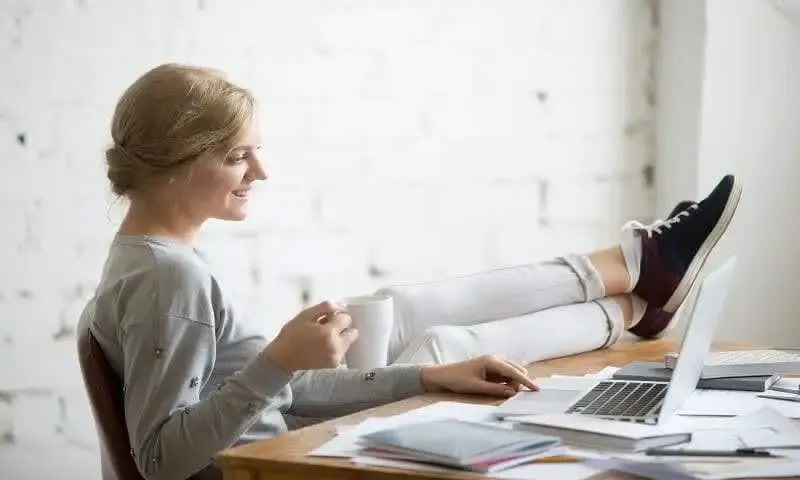 Menina com os pés em cima da mesa, segurando um copo, com cadernos, folhas e um notebook sobre a mesa, enquanto explora o que é SEO.