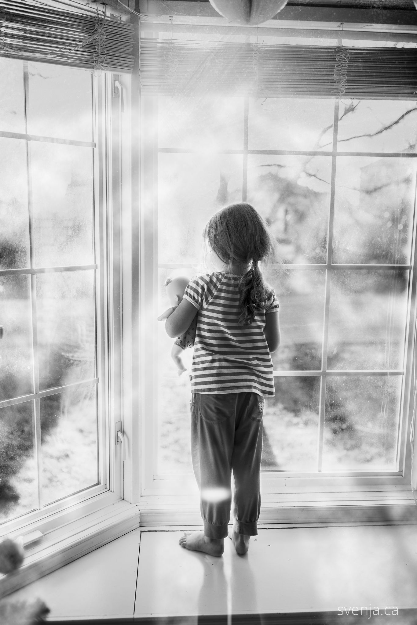 black and white picture of girl looking out the window holding her doll