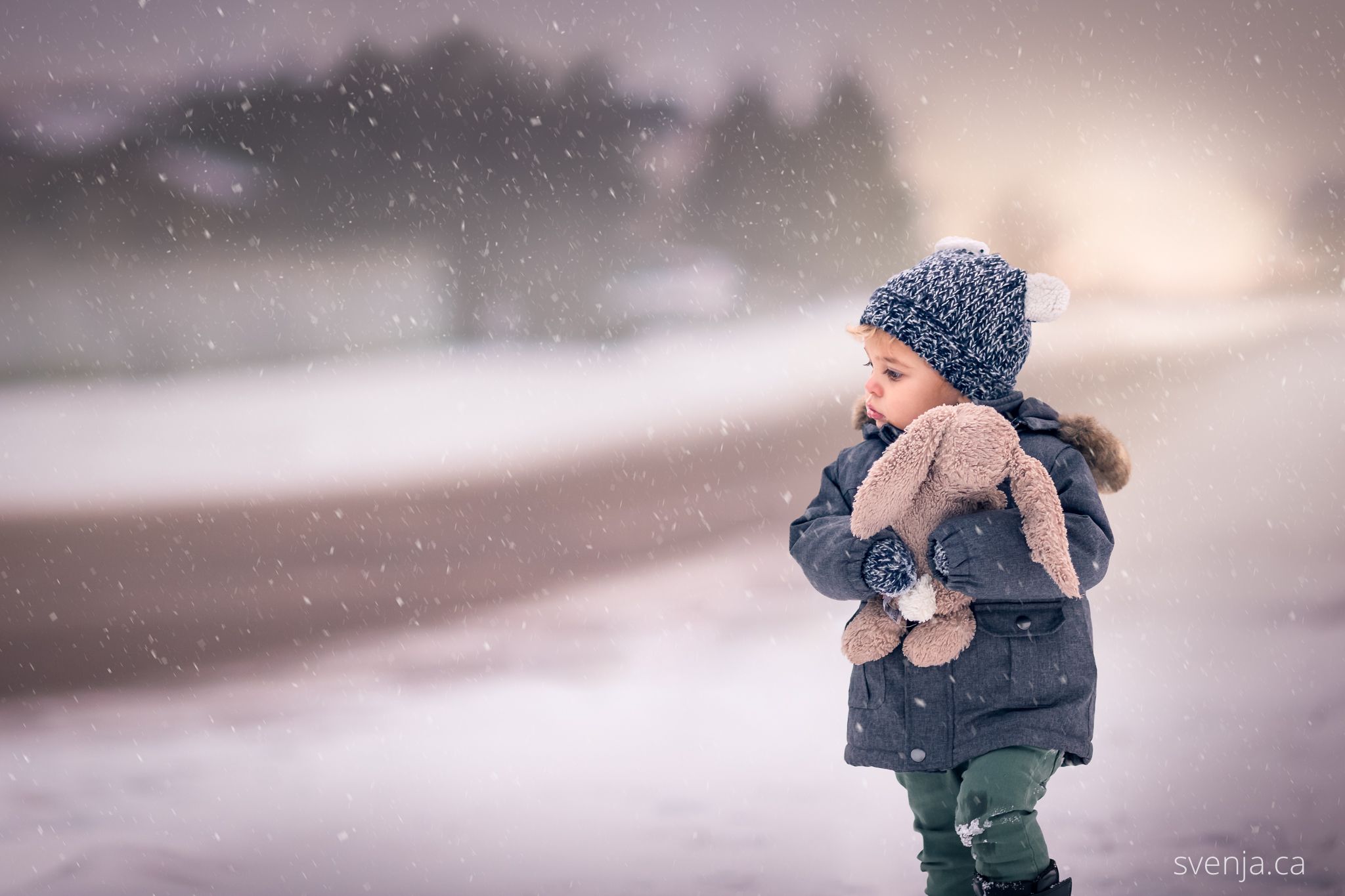 toddler boy in winter coat holds his stuffed bunny with snow on the street