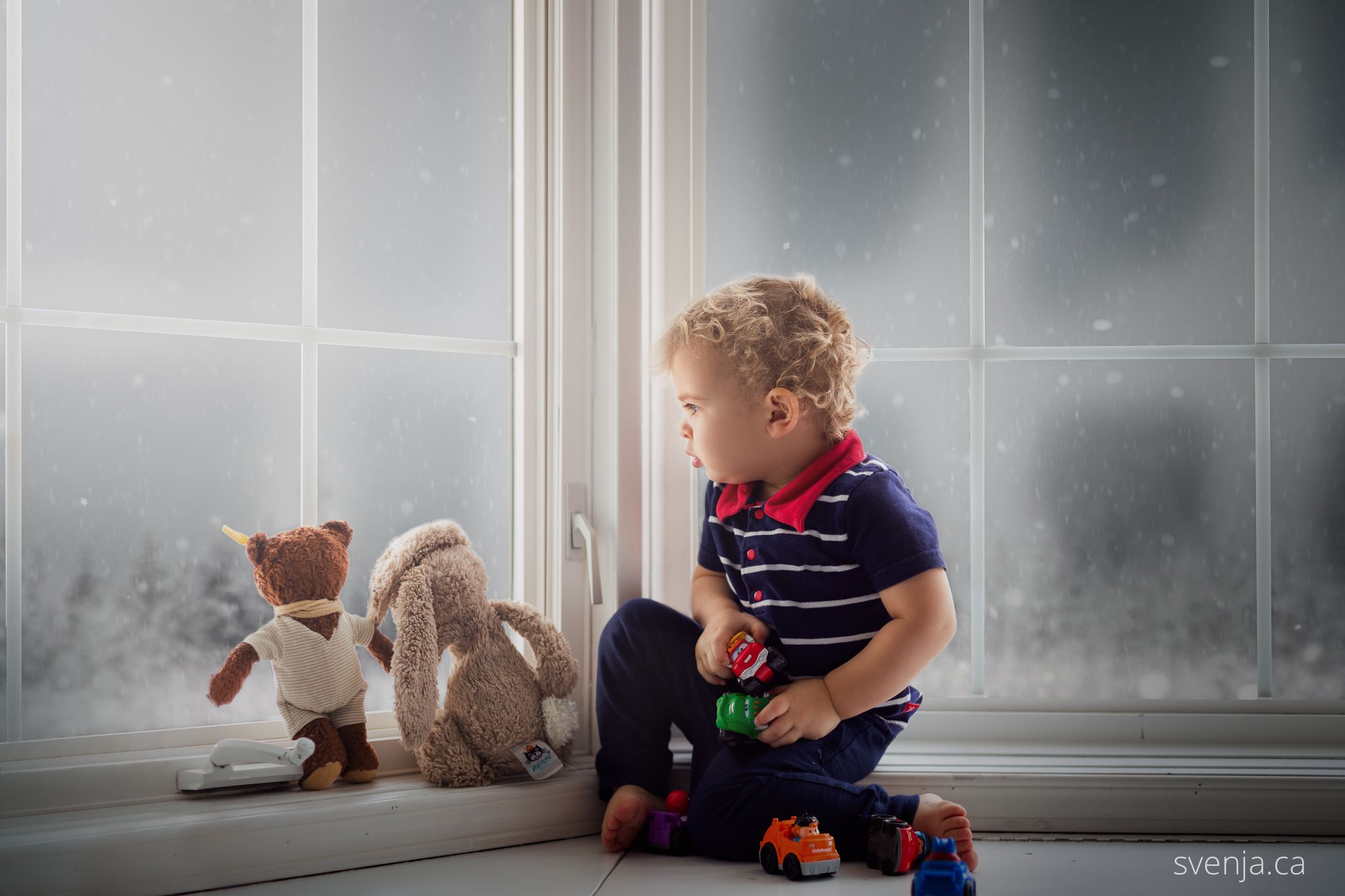 a toddler boy sits with his stuffed animals in a bay window looking at snow