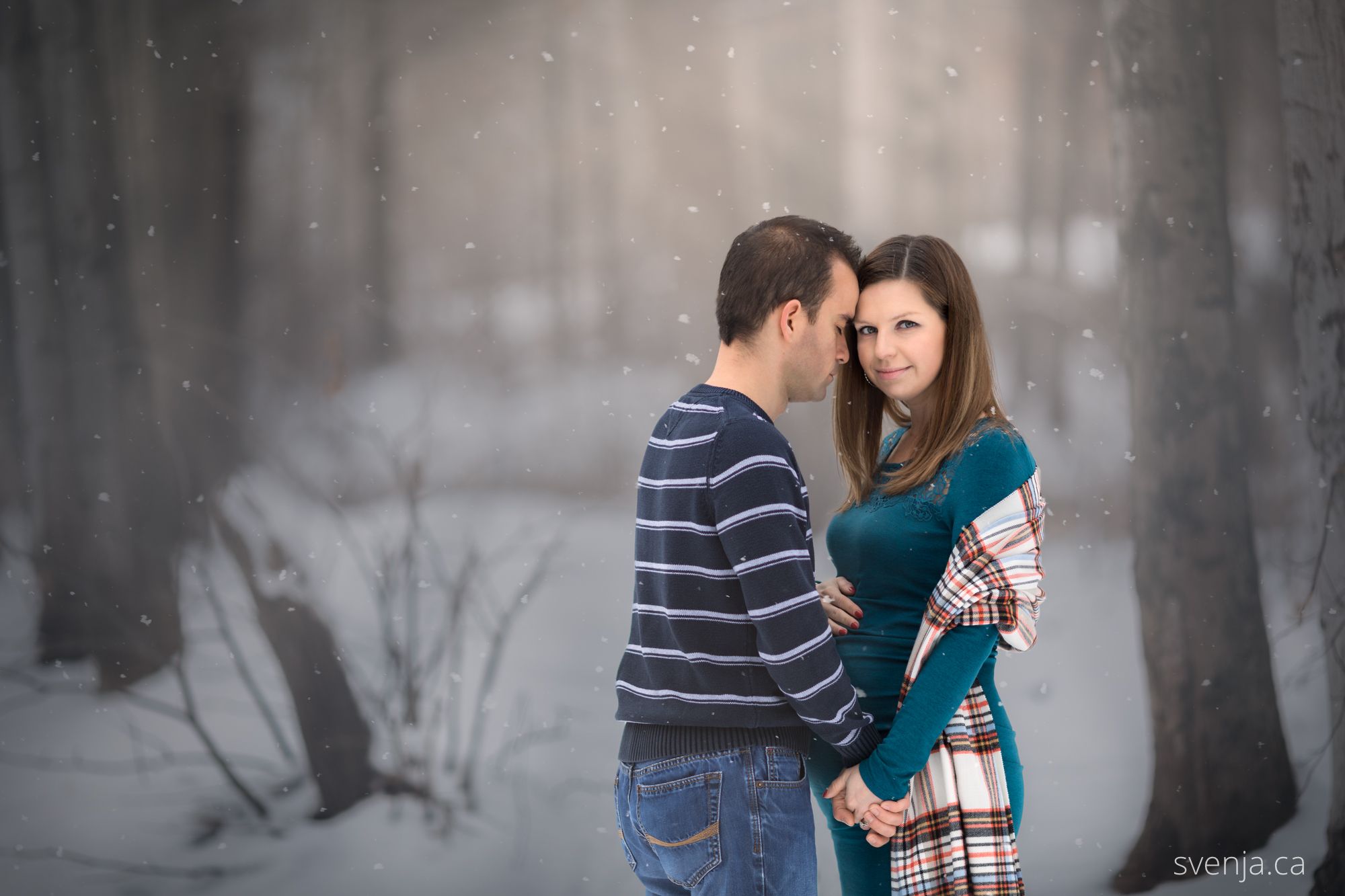 an expecting couple hold each other in a snowy forest