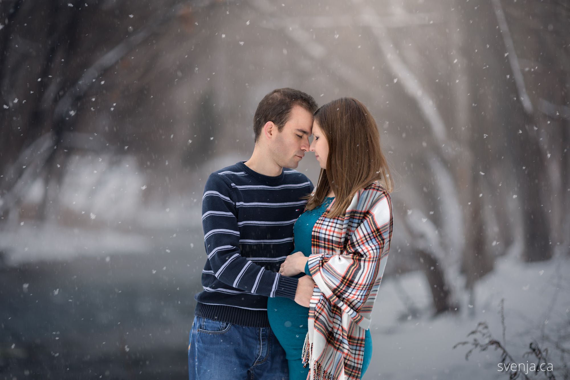 expecting mother and her husband embrace closely with eyes closed with a snowy background