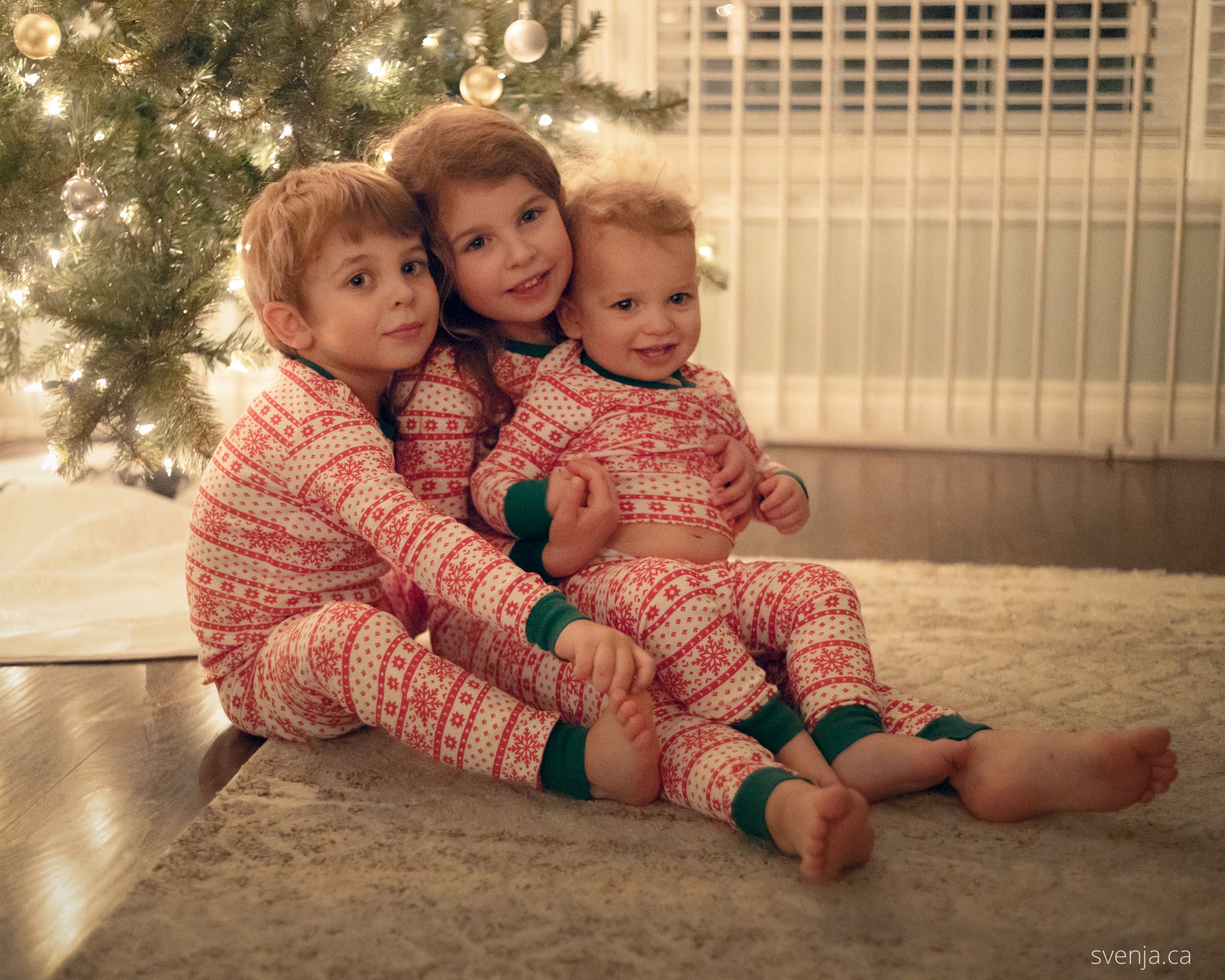 3 children in matching pajamas sit in front of a Christmas tree