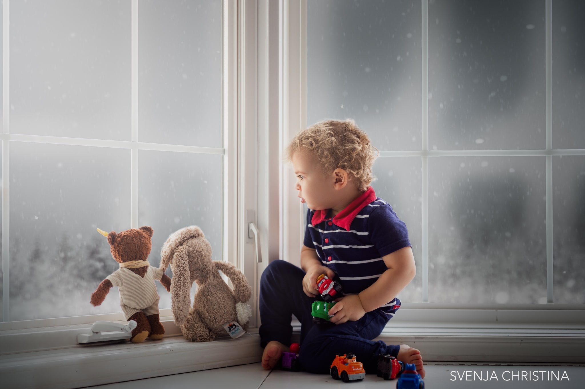 young boy looks outside a window at snow with 2 teddy bears with him