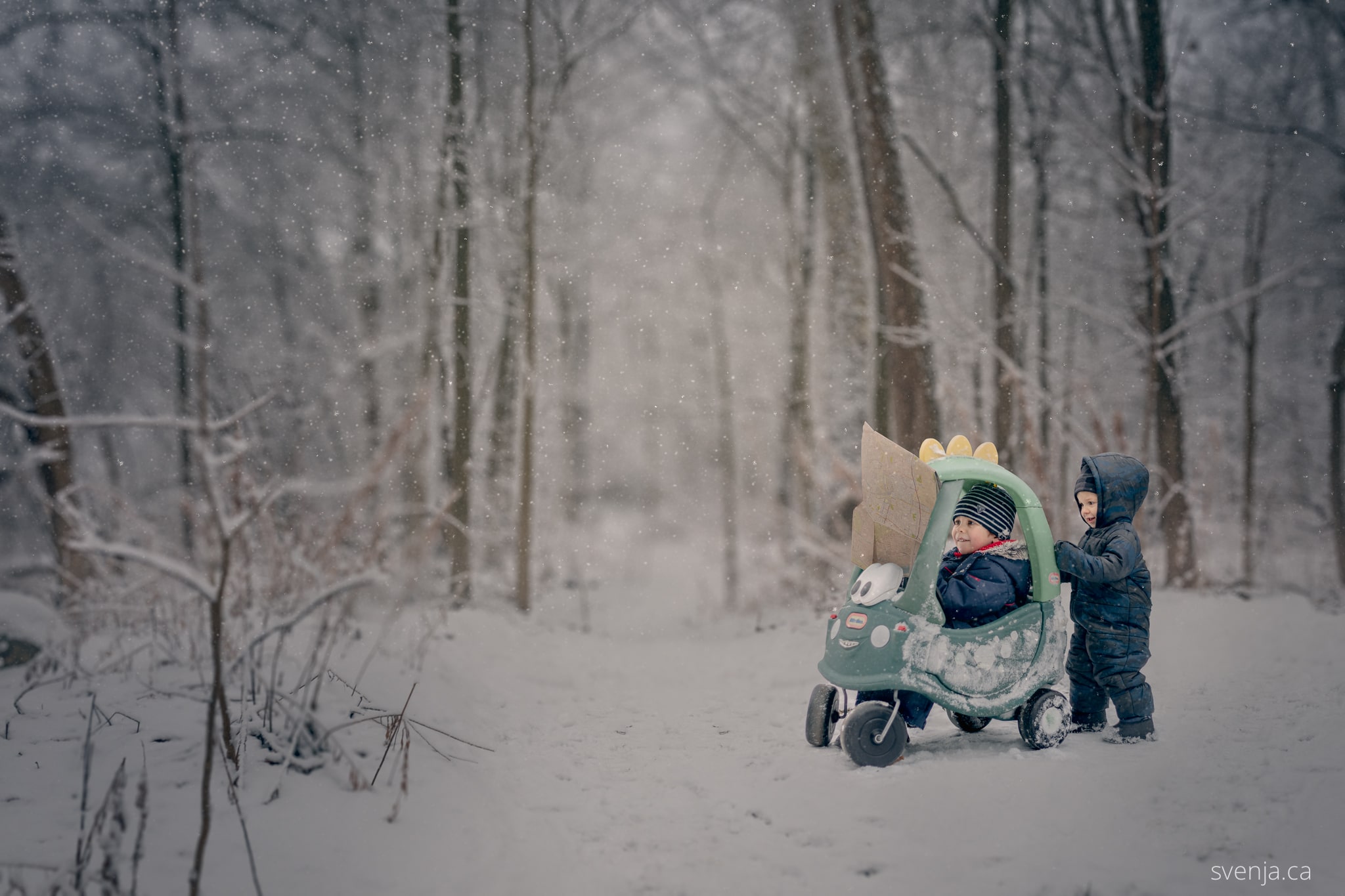 two young boys outside in forest, one sitting in child's toy car, both looking at a map