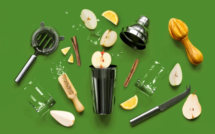 A metal shaker, strainer, ice, a glass and scoop, cinnamon sticks, and various pear and apple slices spilled out for Confetti's Virtual Online Mixology Class