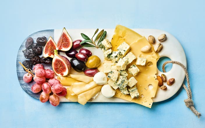 A birds eye view of a white charcuterie board covered in a variety of hard and soft cheeses and fruits for Confetti's Virtual Food Tasting Kits