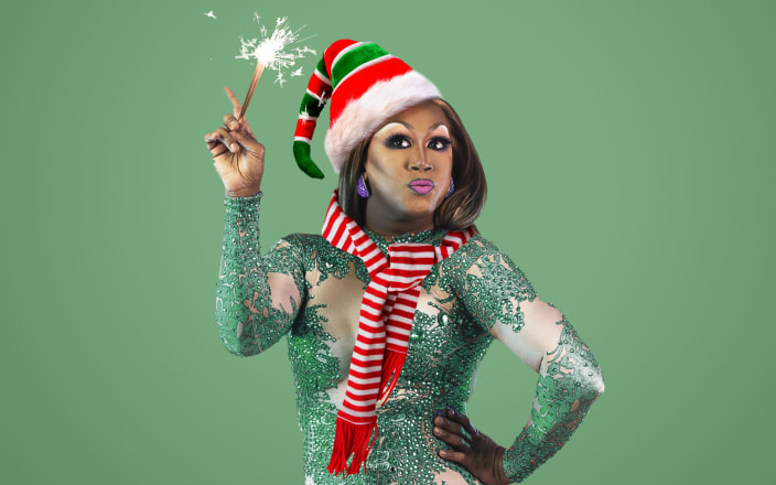 A drag queen wearing an elf hat and scarf, and holding a sparkler for Confetti's Virtual Drag Queen Trivia