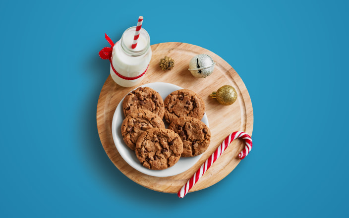 A plate of chocolate chip cookies, a candy cane, and a glass bottle of milk sitting on a wood cutting board with ornaments for Confetti's Virtual Baking Class