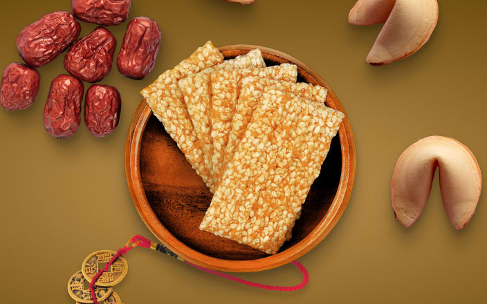 A bowl of sesame crackers next to gold coins, dates, and fortune cookies for Confetti's Asian Snack Box