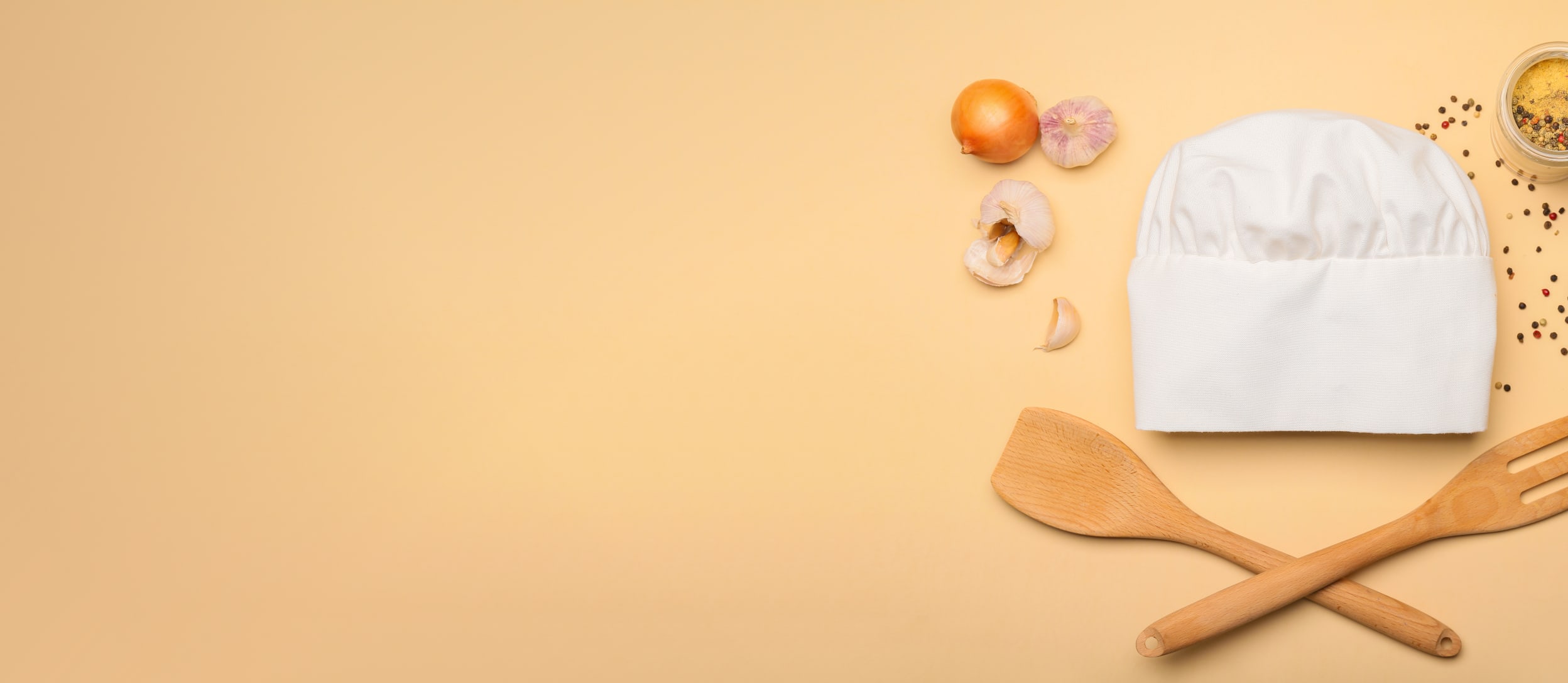 A chefs hat, wooden spatula and fork, onions and garlic, and spilled spices and peppercorns for Confetti's Cooking Classes for Beginners