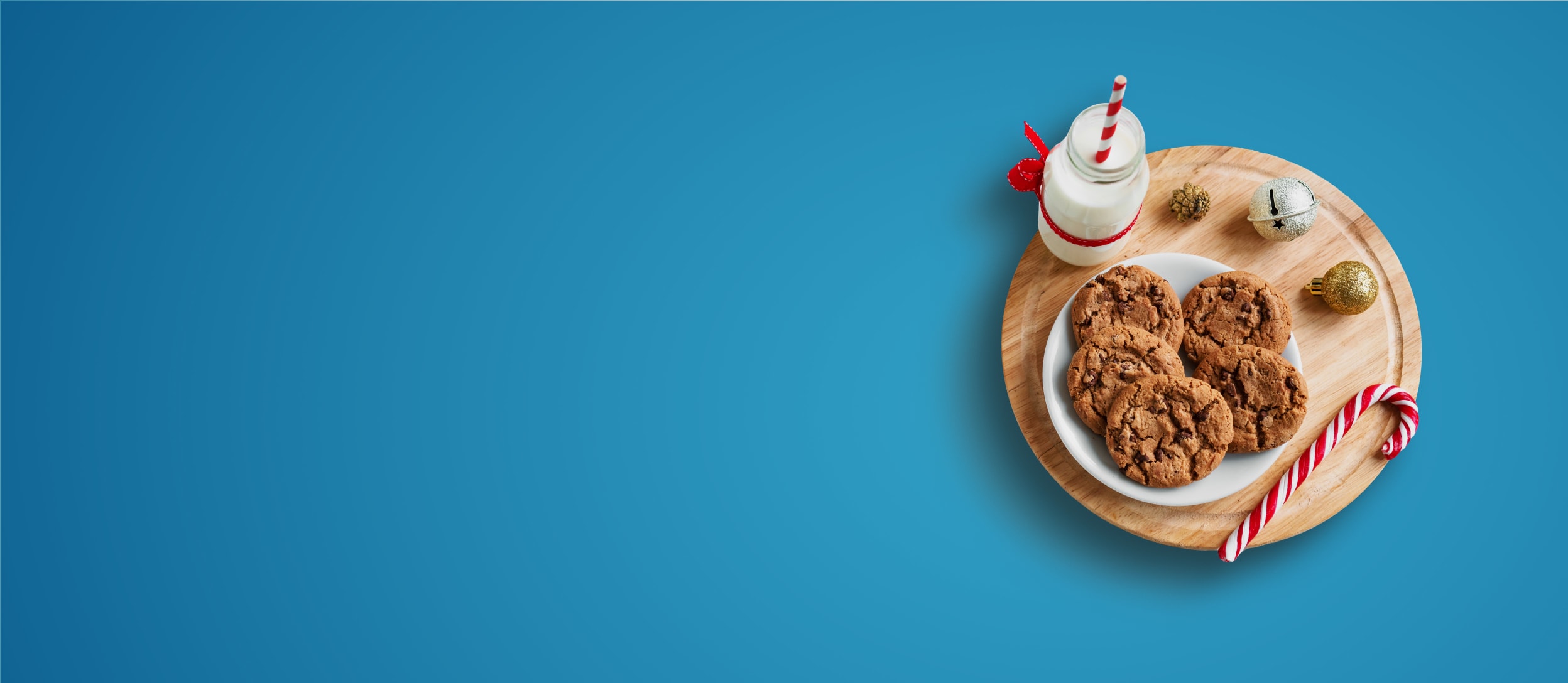 A plate of chocolate chip cookies, a candy cane, and a glass bottle of milk sitting on a wood cutting board with ornaments for Confetti's Virtual Baking Class