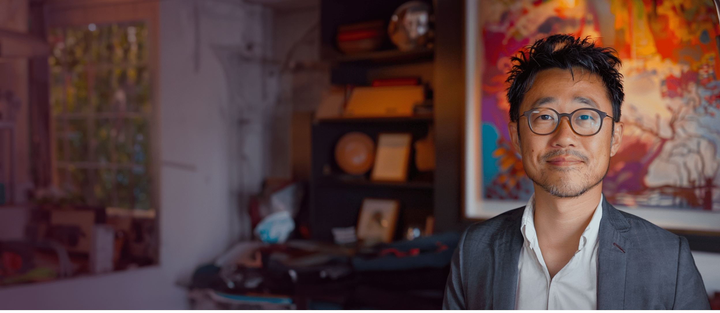 Man dressed in a suit smiling in a home office with a bookshelf and painting (large)