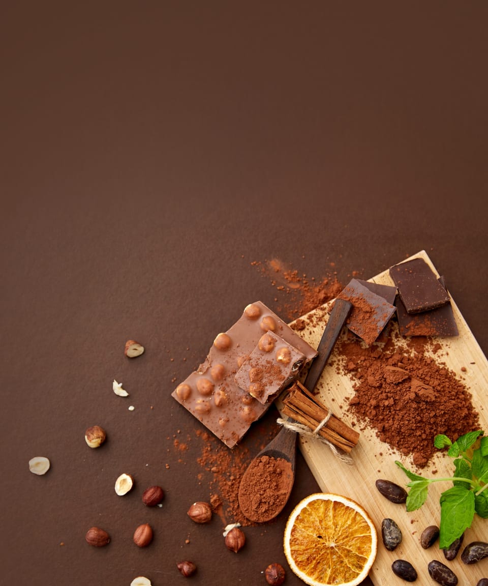 A cutting board with cocoa powder, cinnamon sticks, crushed hazelnuts, and a chocolate bar on it and scattered around for Confetti's Virtual Chocolate Tasting