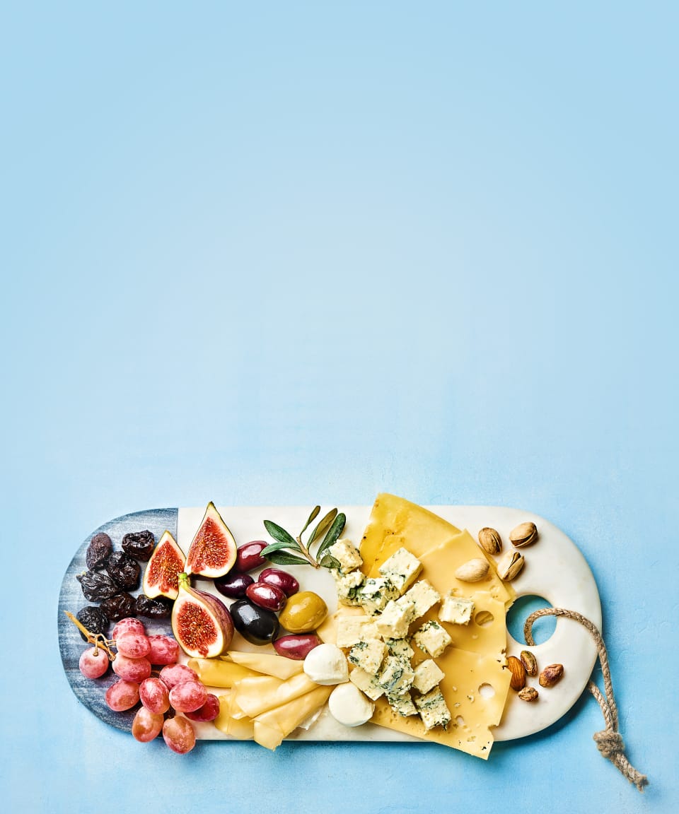 A birds eye view of a white charcuterie board covered in a variety of hard and soft cheeses and fruits for Confetti's Virtual Cheese Tasting